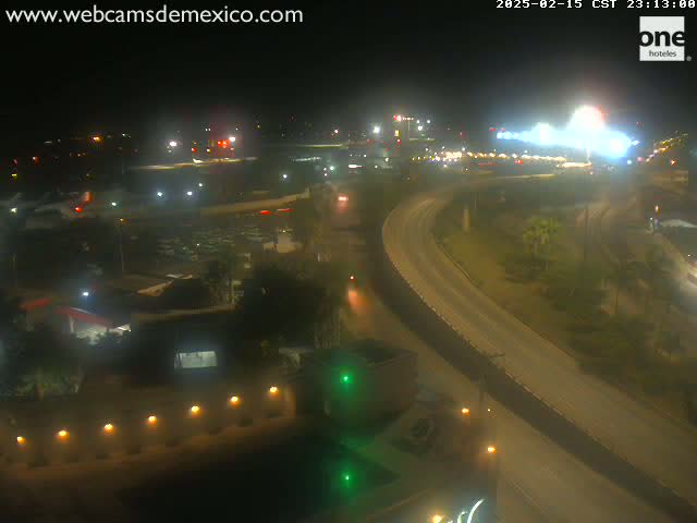 A nighttime, high-angle view of a highway curving through a city with surrounding buildings and parking lots illuminated by lights.