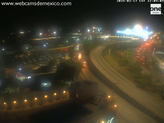 A nighttime, high-angle view shows a highway with moderate traffic curving through a landscape with sparse vegetation and buildings, under dark conditions.