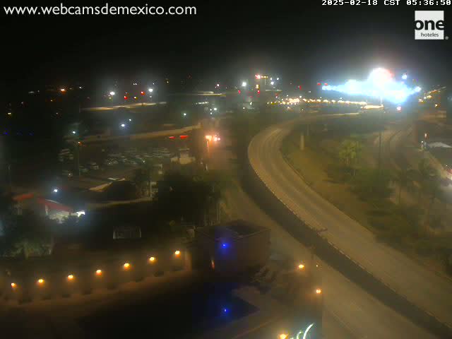 A nighttime, high-angle view shows a curving roadway with sparse traffic, illuminated buildings, and surrounding vegetation.