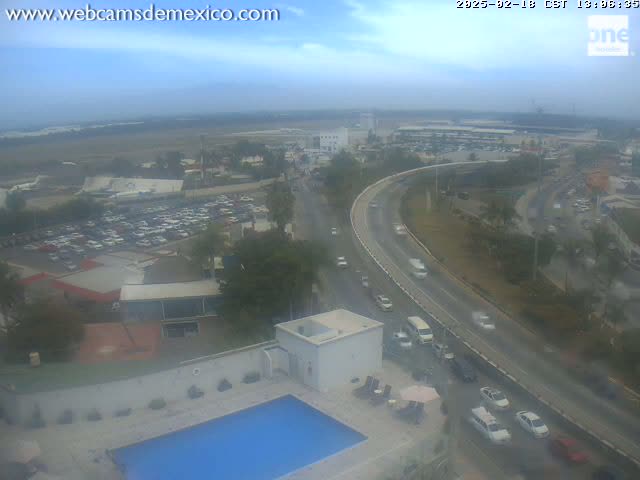 An aerial view shows a highway curving around a large parking lot and buildings near an airport under an overcast sky.