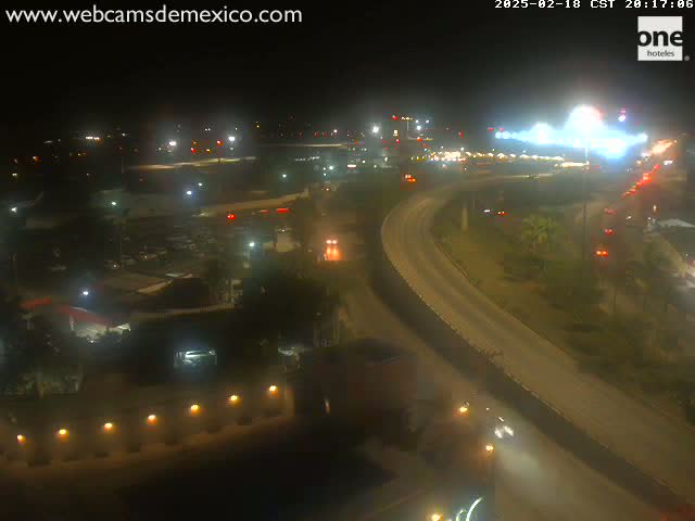 An elevated view at night shows a highway curving through a landscape with sparse vegetation and buildings, illuminated by artificial lights.