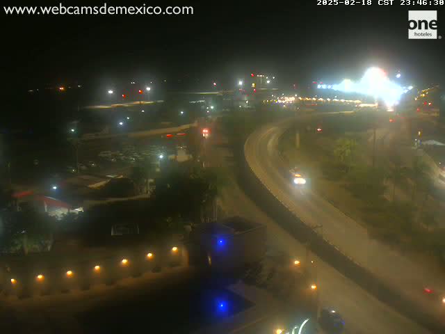 A nighttime, slightly hazy view of a highway curving around a building complex with scattered lights and a few vehicles.