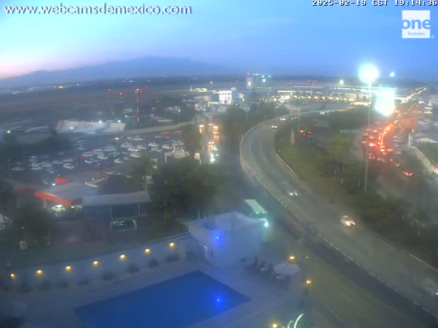 An elevated view at twilight shows a highway curving through an airport area with parking lots, buildings, and a swimming pool at a hotel, under a clear sky.
