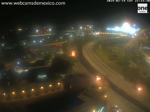 A nighttime aerial view shows a highway curving through a city with dimly lit buildings and sparse vegetation under a clear sky.