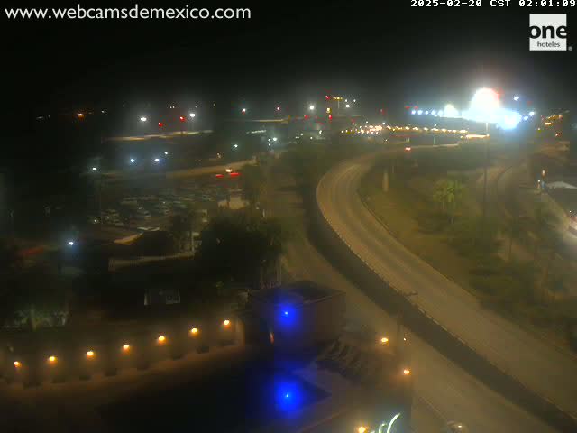 A nighttime, high-angle view shows a highway curving through a sparsely lit landscape, with buildings and parking lots visible in the background.
