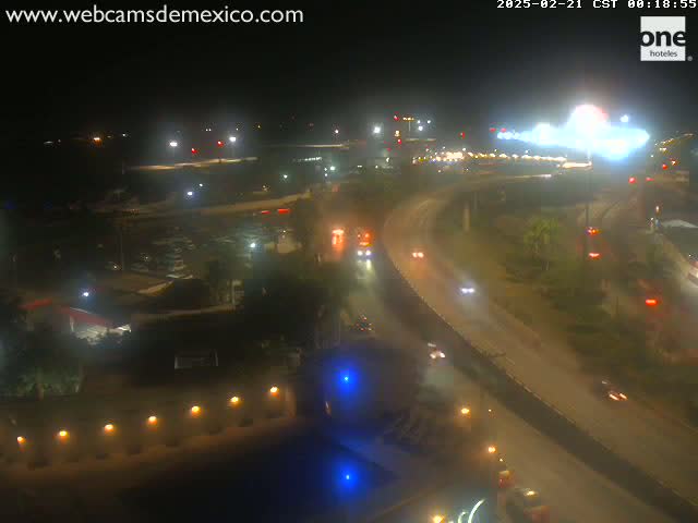 A nighttime, wet, overhead view of a highway curving through a city with sparse vehicle traffic and surrounding buildings faintly lit.