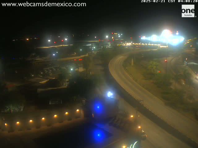 A nighttime aerial view of a highway curving through a well-lit area with buildings and sparse vegetation.