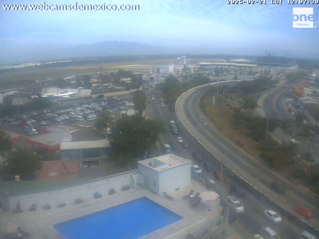 An elevated view on an overcast day shows a highway curving past a building with a swimming pool, parking lots, and an airport in the distance.