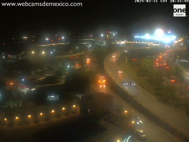 A nighttime aerial view shows a highway interchange near an airport with cars driving on the road and planes on the tarmac under mostly clear skies.
