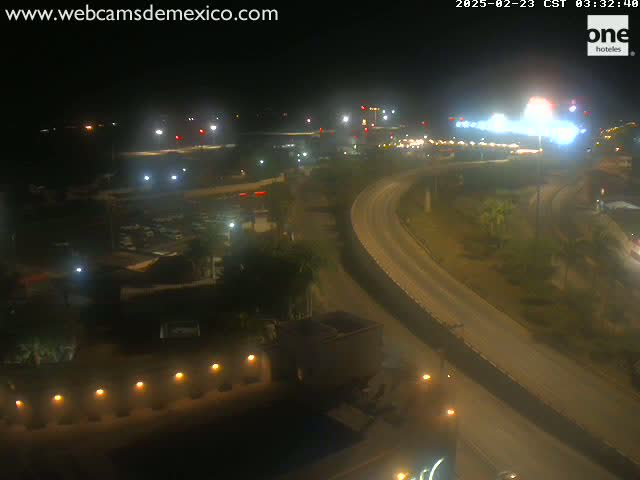 A nighttime aerial view of a curving roadway, illuminated by streetlights, with sparse vegetation and buildings visible in the surrounding area under clear skies.
