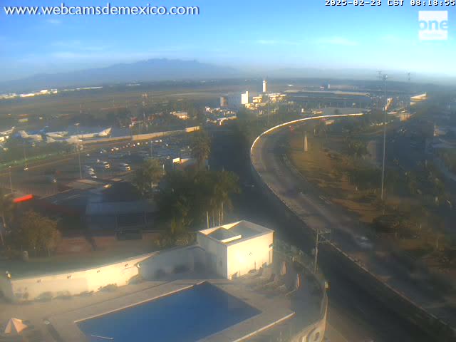 An elevated view shows a large roadway curving around a complex of buildings near an airport under a hazy, sunny sky with distant mountains visible on the horizon.