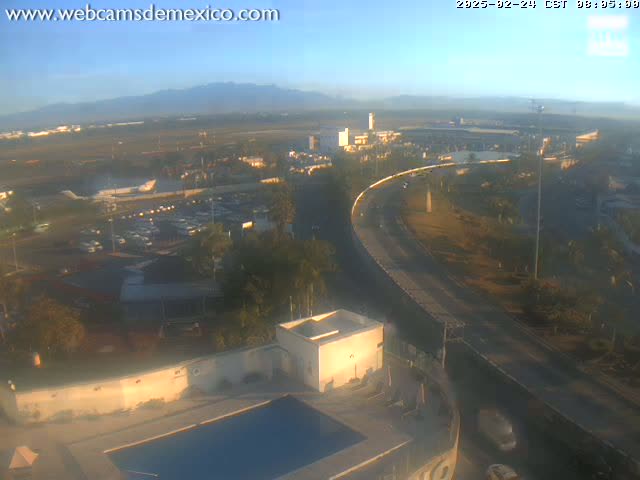 A high-angle, long shot on a clear, sunny day shows a highway curving around a building complex with a pool, near an airport with distant mountains.