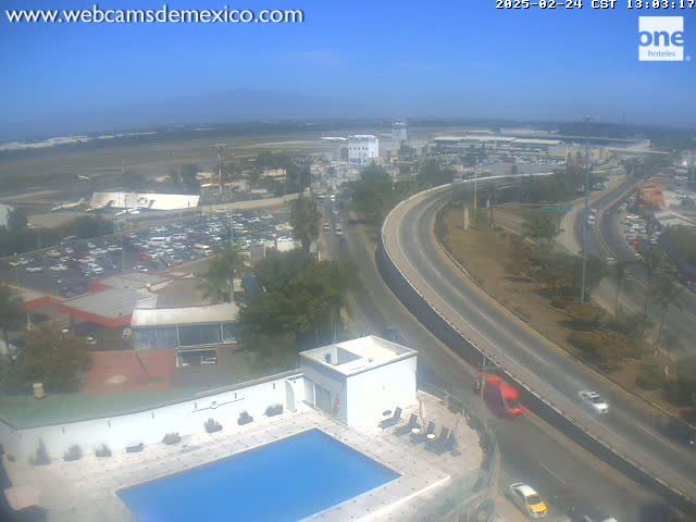 A high-angle, long shot on a sunny day shows a swimming pool next to a building overlooking a highway curving around a large parking lot and airport in the distance.