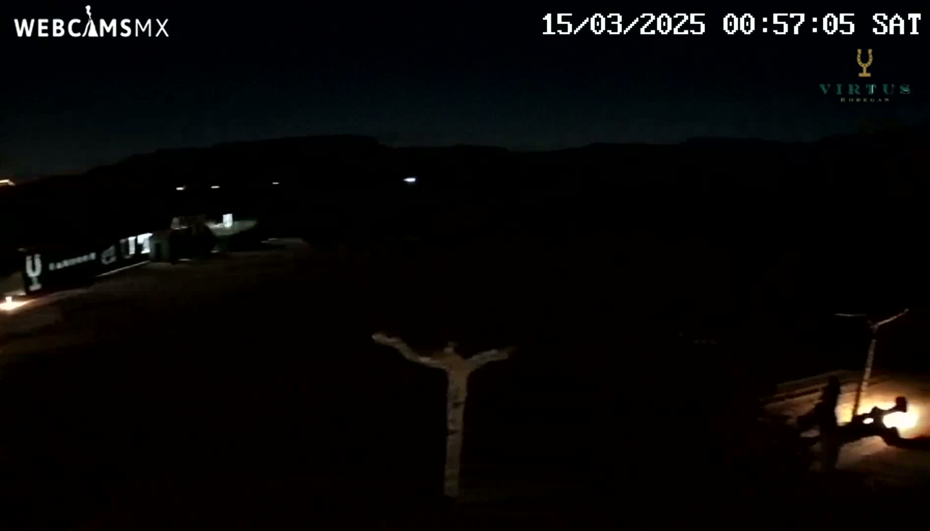 A dark, nighttime scene shows a mostly obscured area with a few faint lights and what appears to be a person or small group of people sitting near a light source in the lower right corner.