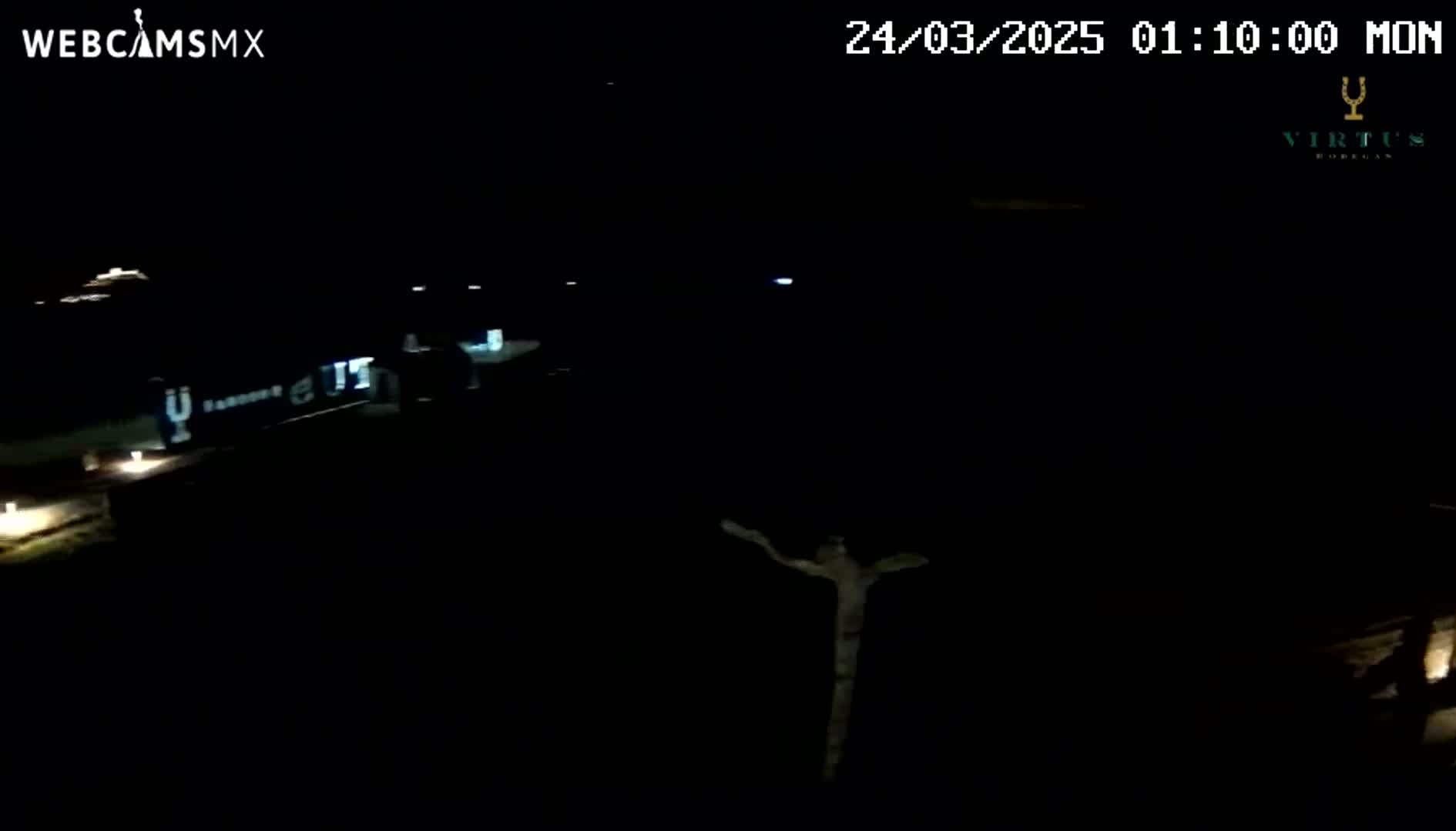 A nighttime, outdoor view shows a dark area with a few distant lights and a vaguely crucifix-shaped object in the center.