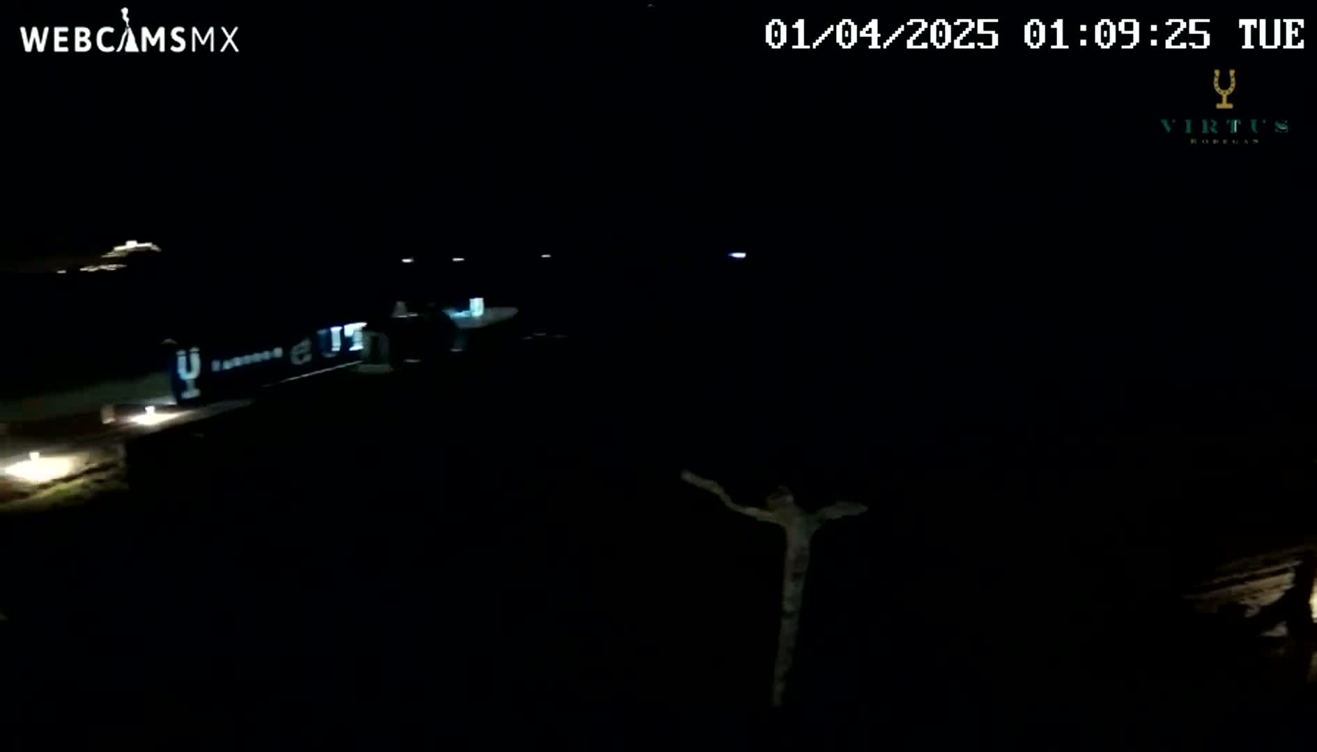 A dark nighttime scene shows a long, light-colored object, possibly a vehicle, with faint lights, and a vaguely cross-shaped object in the foreground.