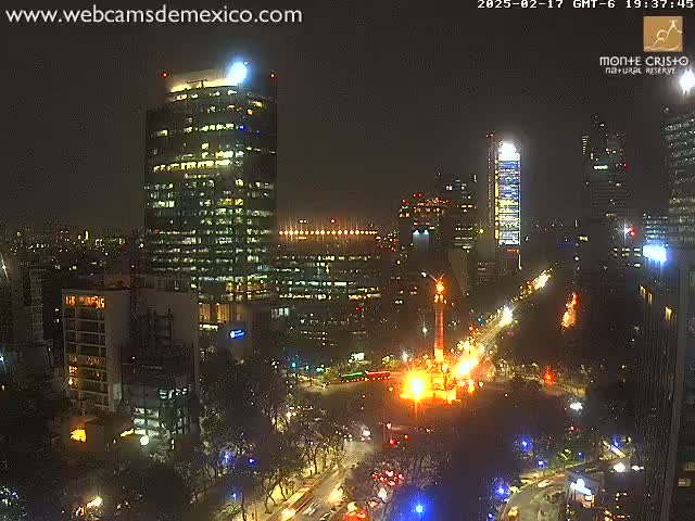 A nighttime cityscape view shows illuminated skyscrapers and city streets, with a monument brightly lit at a street intersection.