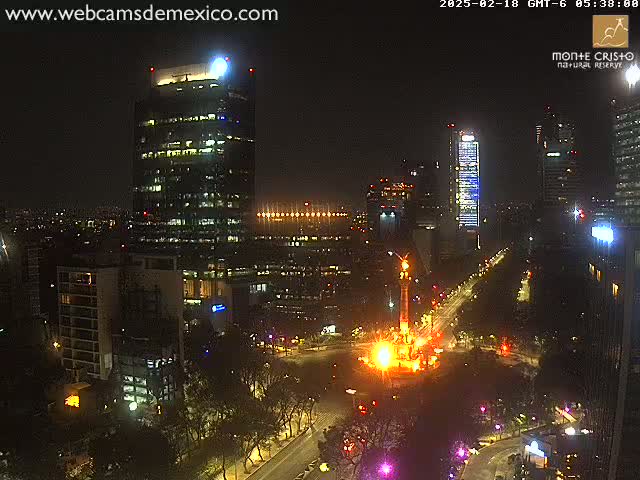 A nighttime city scene shows illuminated skyscrapers and a brightly lit monument in a large plaza with surrounding streets.