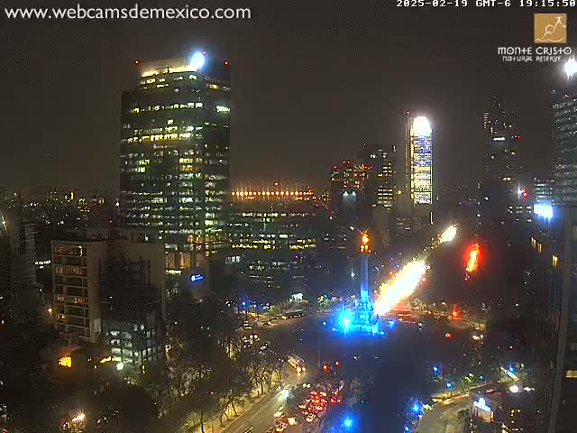 A nighttime city scene shows a tall building illuminated, with a large monument in a plaza displaying bright blue and orange lights, surrounded by city streets and other buildings.
