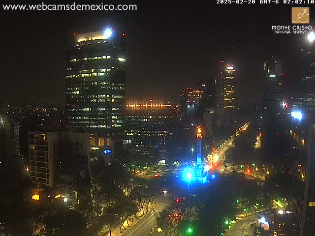 A nighttime, high-angle view of a city illuminated with lights, featuring a tall building in the foreground and a monument lit in blue at the center of a large intersection.