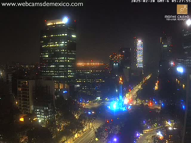 A nighttime aerial view of a city's illuminated cityscape, featuring several skyscrapers and a brightly lit monument in a plaza.