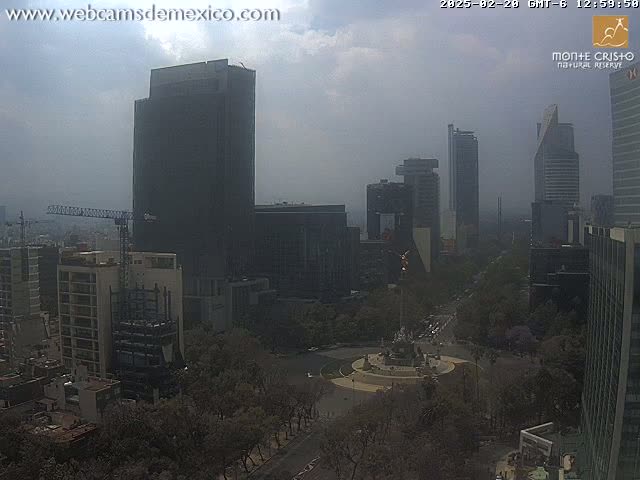 A hazy, partly cloudy day reveals a cityscape featuring numerous skyscrapers and a large monument in a circular plaza surrounded by trees.