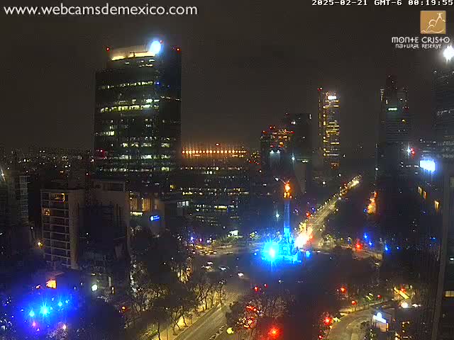 A nighttime aerial view of a city with illuminated skyscrapers and a monument, under a cloudy sky.