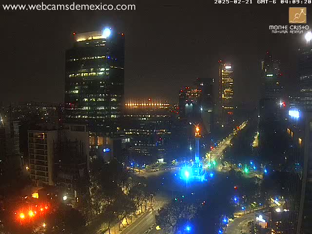 A nighttime, city view shows illuminated skyscrapers and a monument in a plaza, with streets and trees visible under clear skies.