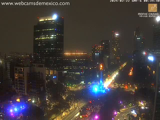 A nighttime aerial view of a city with illuminated skyscrapers and a brightly lit monument at a large intersection.