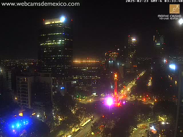 A nighttime cityscape view shows brightly lit buildings surrounding a monument illuminated in pink and purple light, under clear skies.