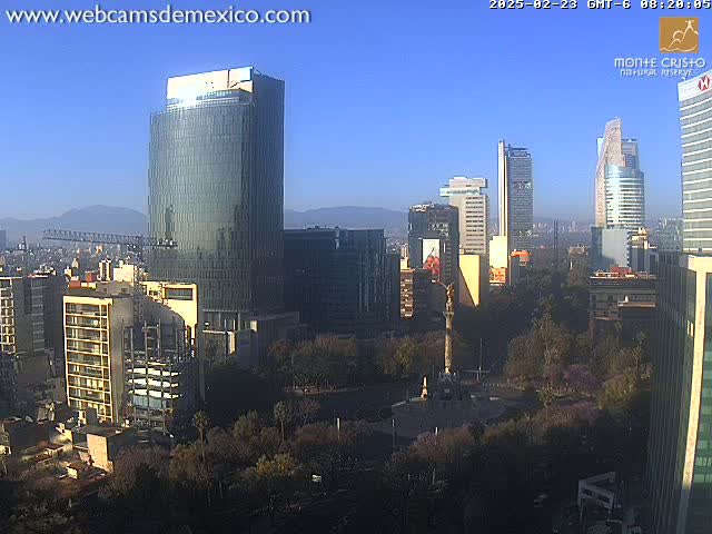 A city skyline with several tall buildings, a monument in a plaza, and surrounding trees under a clear blue sky.