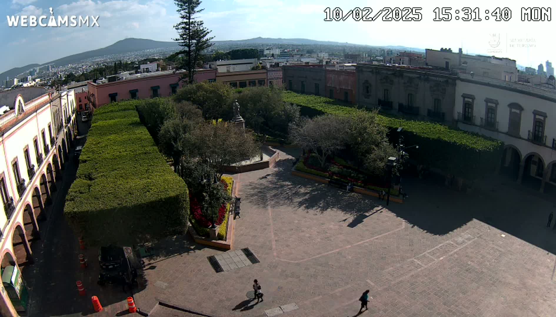 A sunny view of a town square with neatly trimmed hedges, colonial-style buildings, a statue in a plaza, and a few pedestrians.