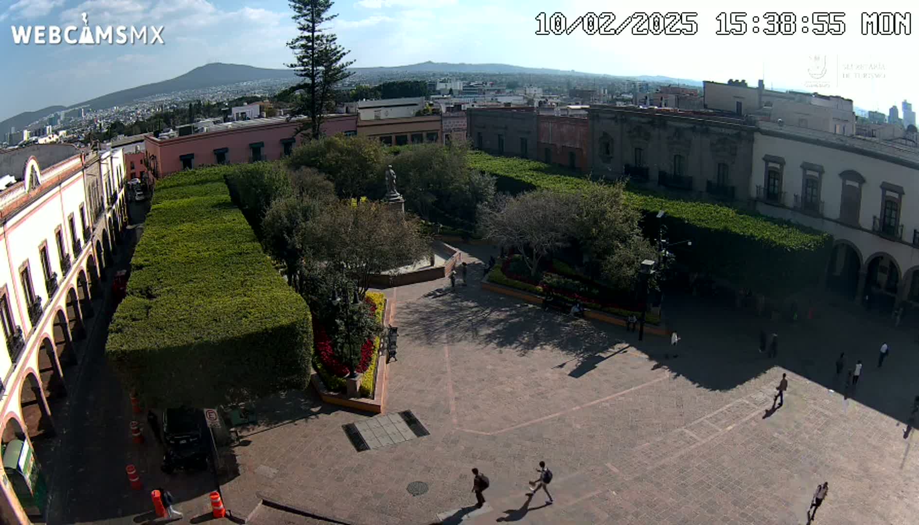 A sunny day overlooking a town square with a central plaza surrounded by buildings and hedges, a few people walking in the square.