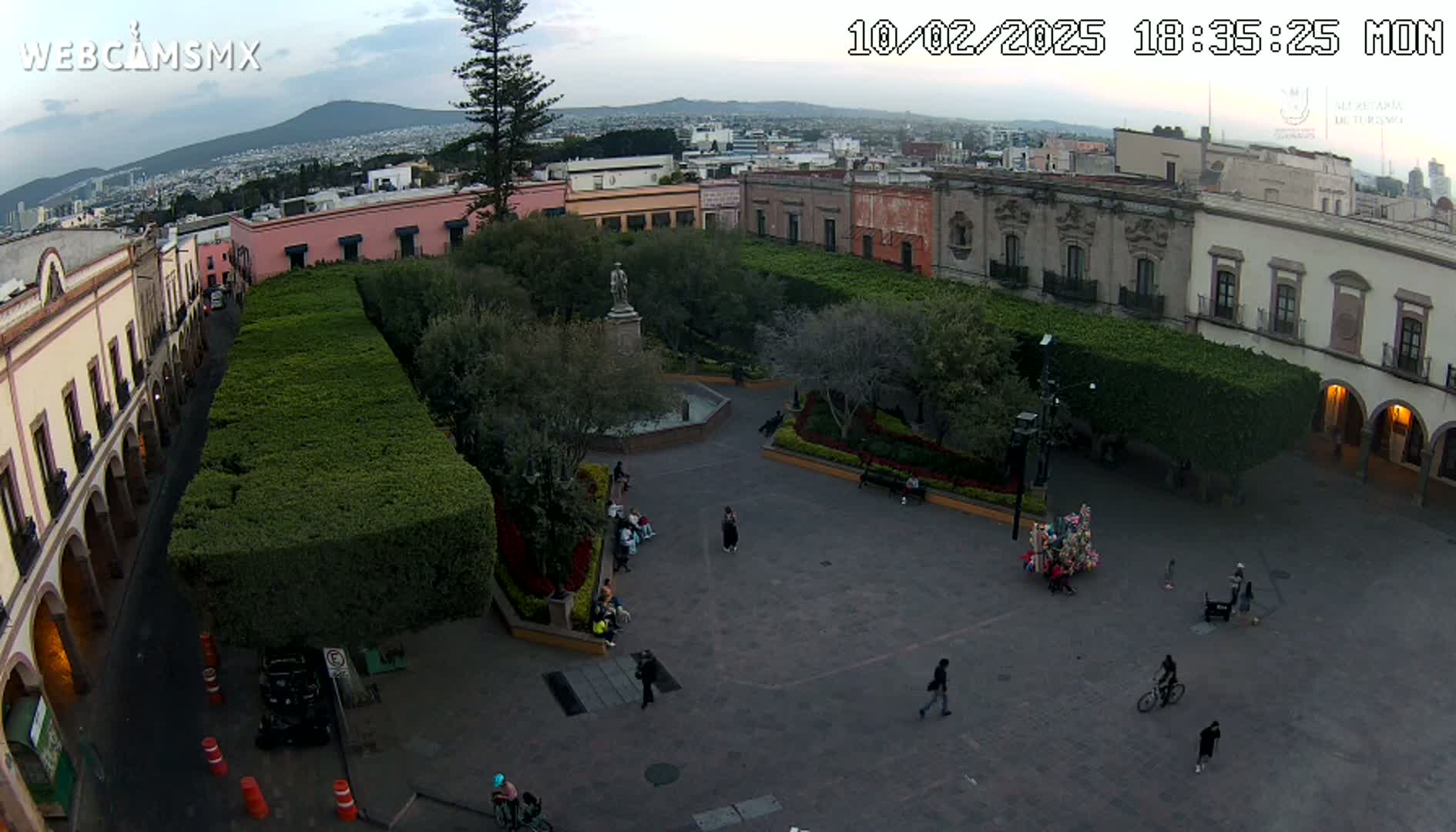 An aerial view of a town square with a central plaza, surrounded by colonial-style buildings under a partly cloudy sky.