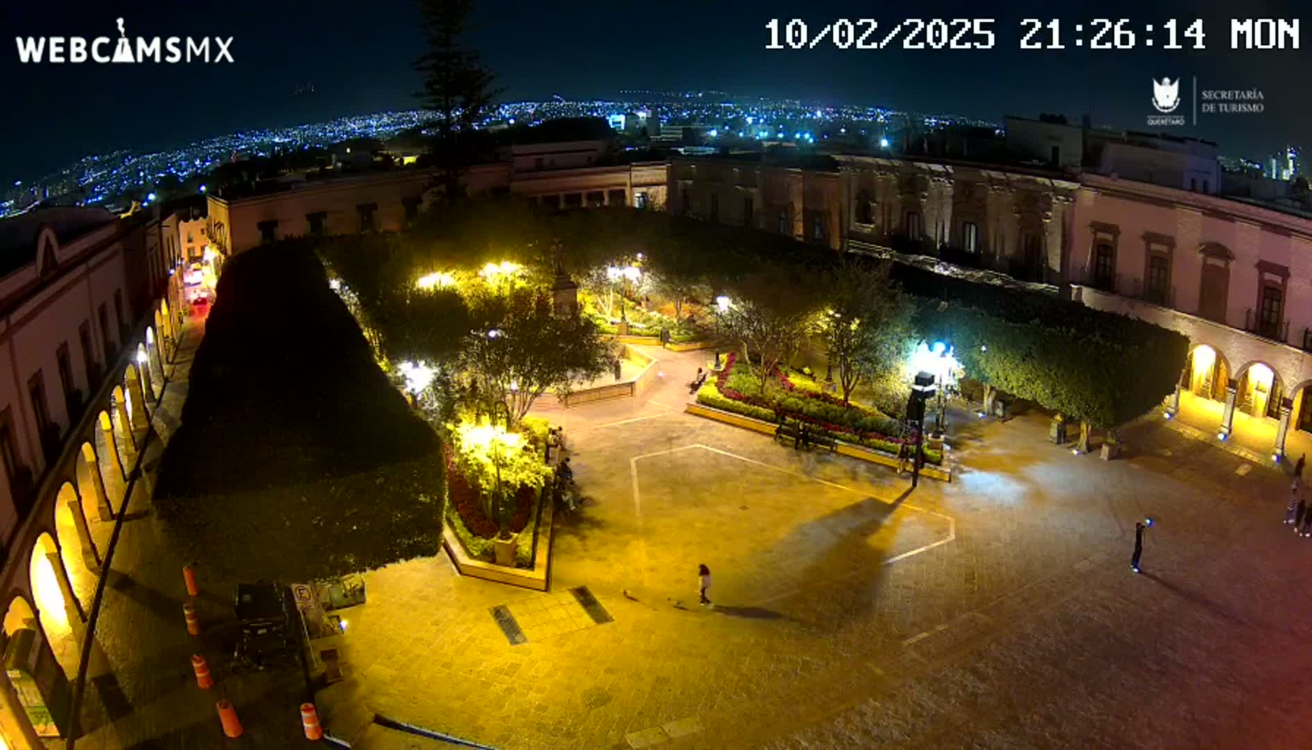 A nighttime, illuminated view of a town square with surrounding buildings, landscaping, and a few people present.