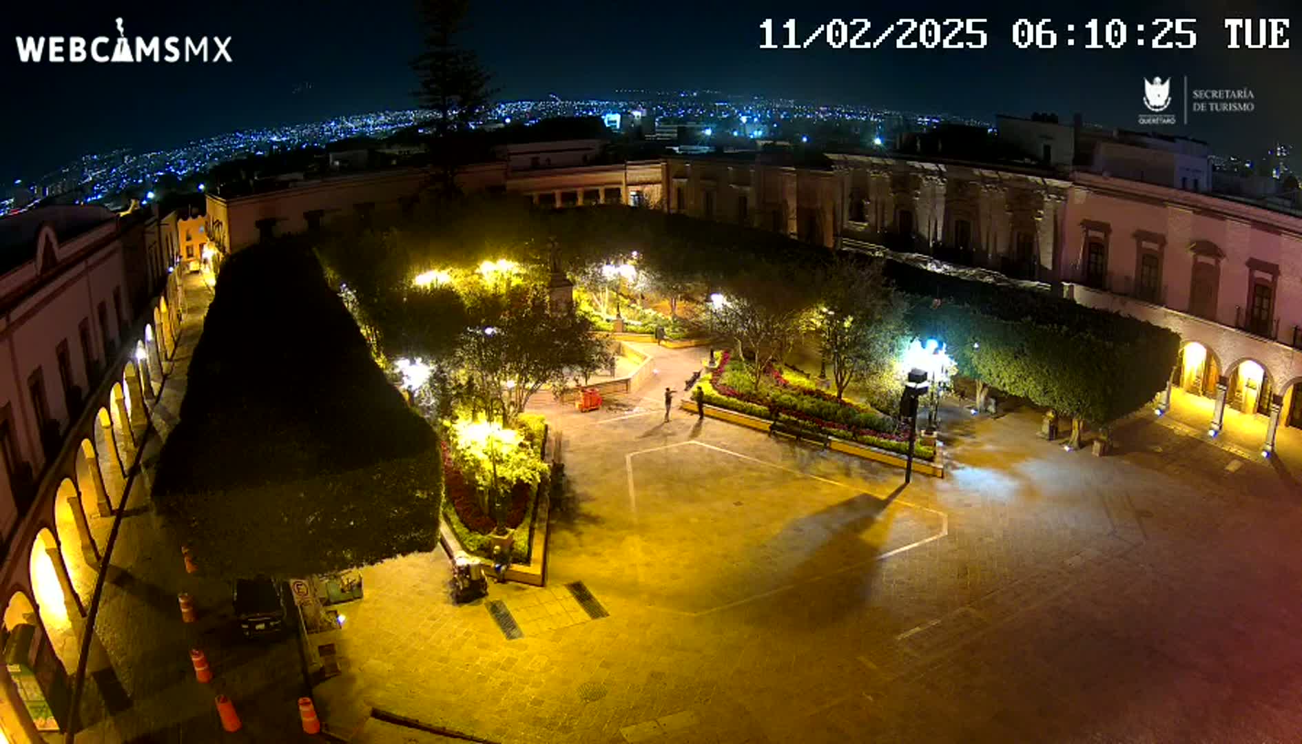 A nighttime aerial view of a brightly lit plaza surrounded by colonial-style buildings, with a city skyline visible in the background.
