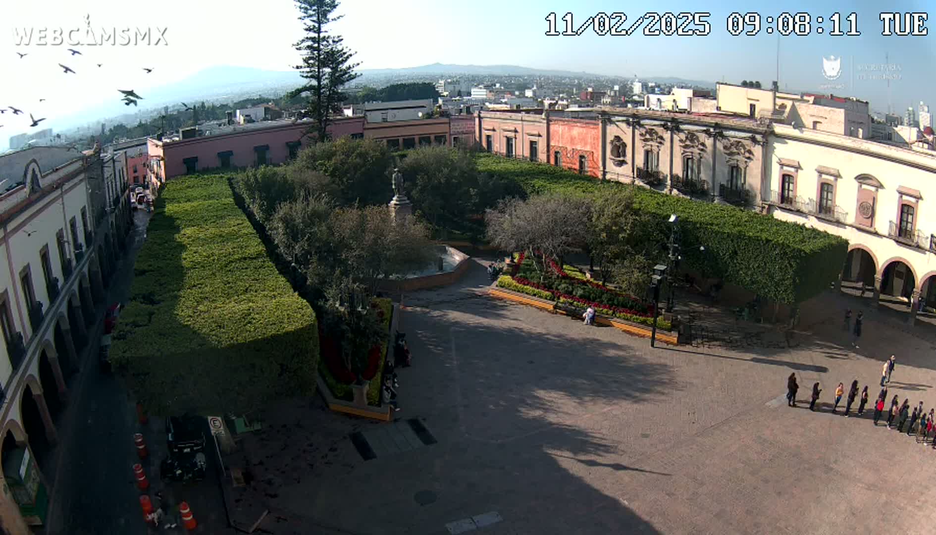 A high-angle, sunny view shows a town square with a central fountain, surrounded by manicured hedges and colonial-style buildings.