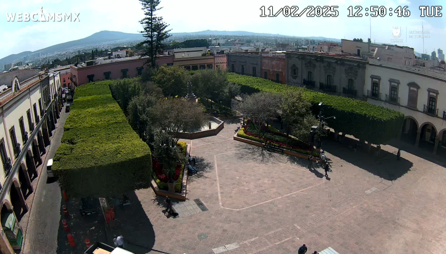 An aerial view of a town square with a central fountain, surrounded by meticulously trimmed hedges, colonial-style buildings, and a distant mountain under sunny skies.