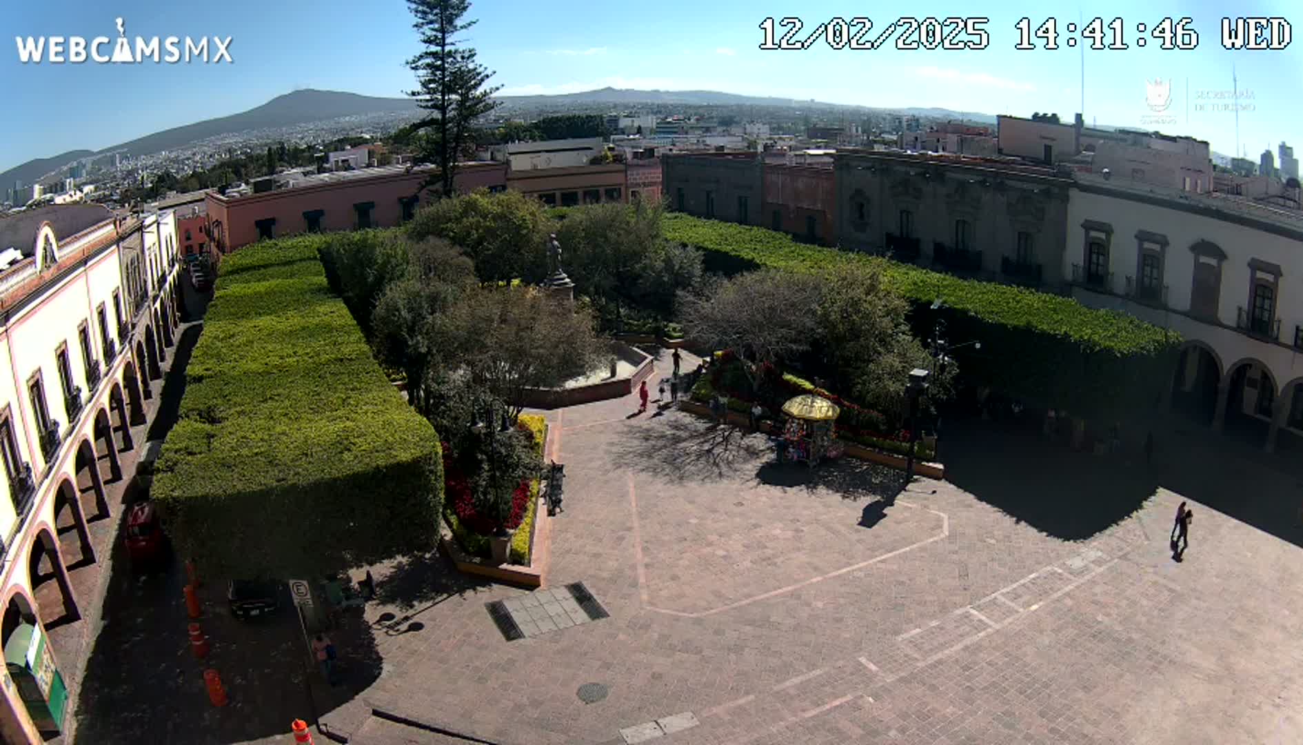 A sunny aerial view of a plaza surrounded by colonial-style buildings and neatly trimmed hedges, with a few people scattered throughout.