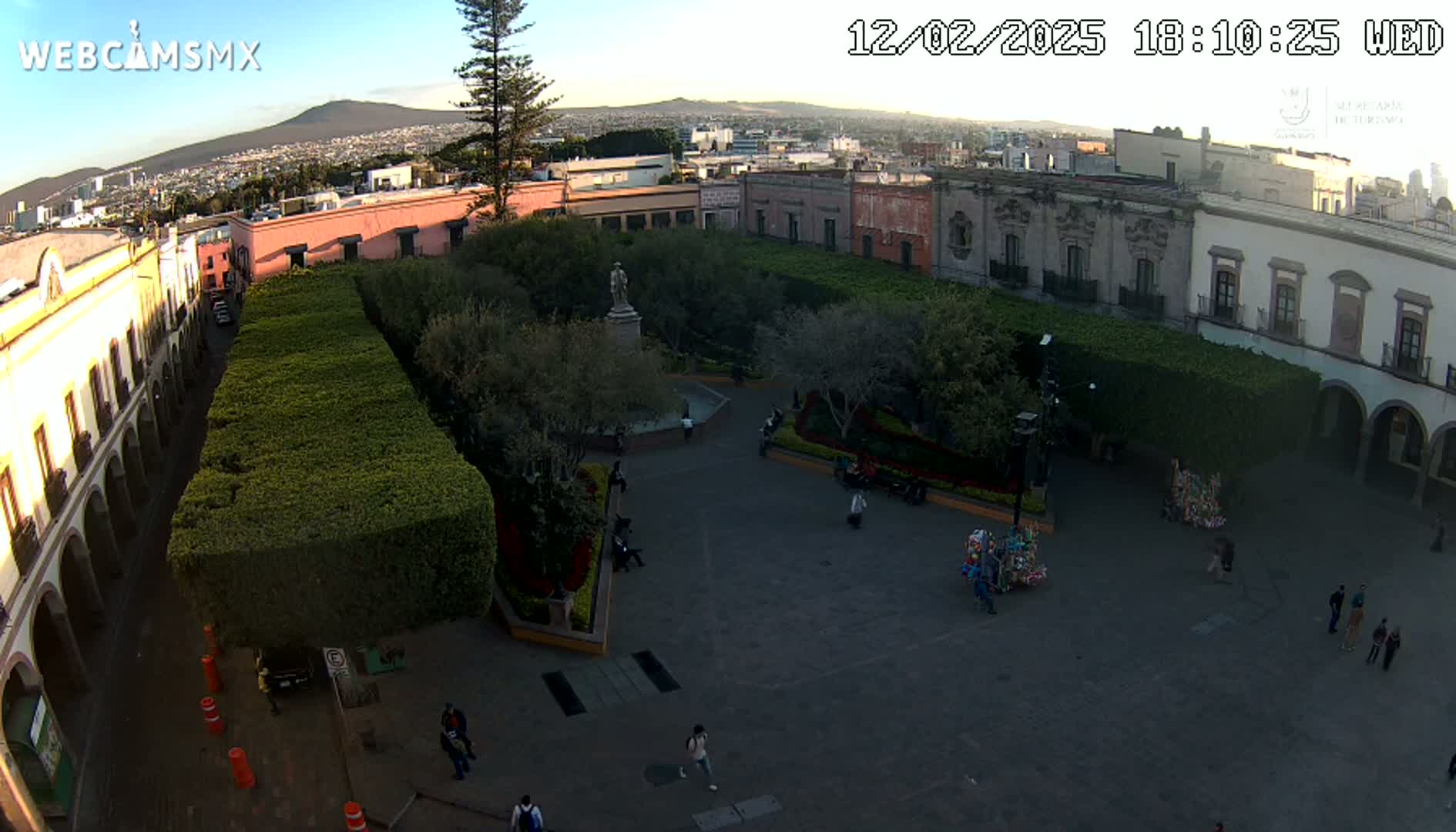 An aerial view shows a town square with a statue in the center, surrounded by neatly trimmed hedges and colonial-style buildings, under a clear evening sky.