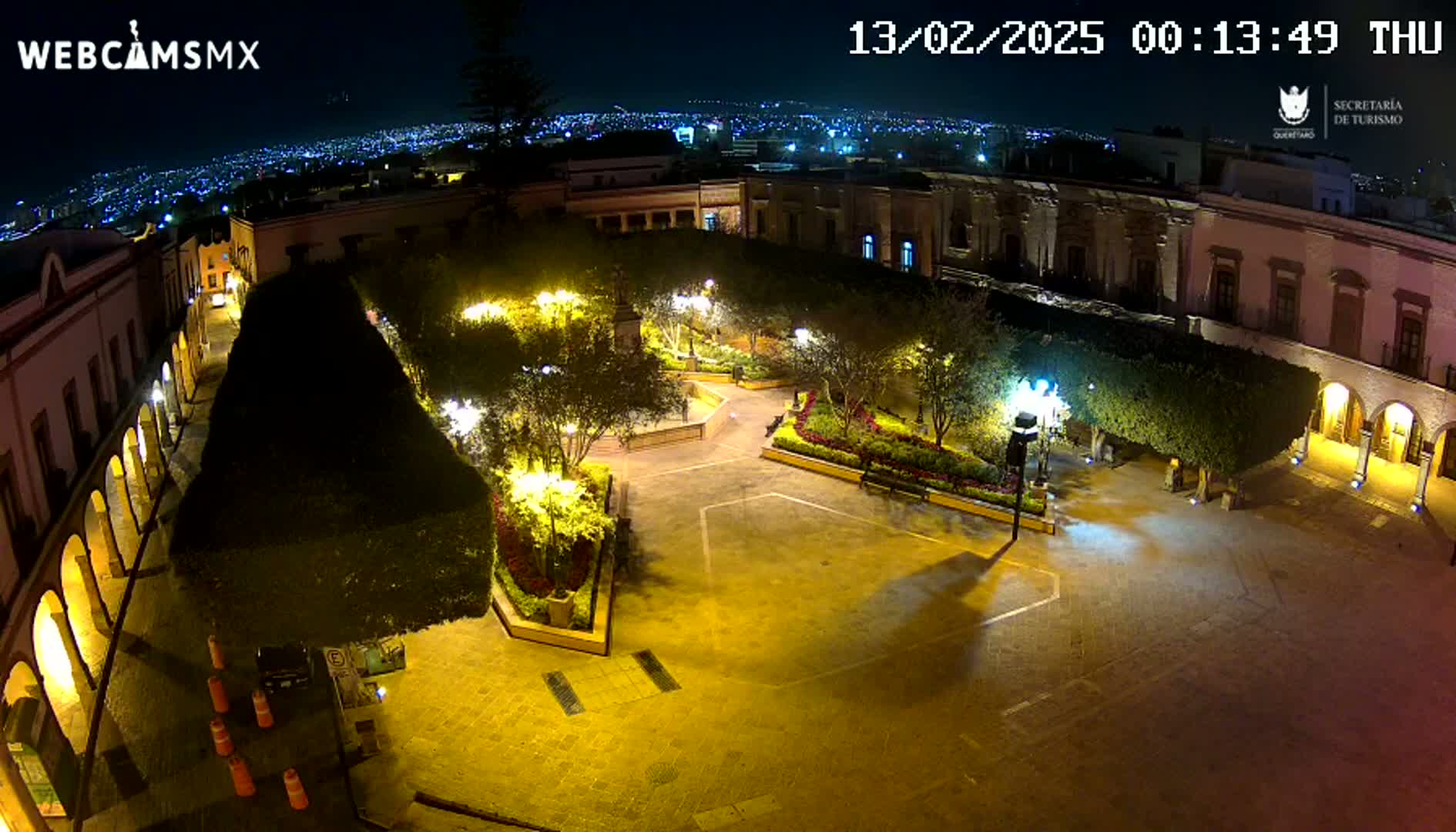 A nighttime, illuminated plaza surrounded by buildings, featuring landscaped gardens and trees, overlooks a distant city skyline.
