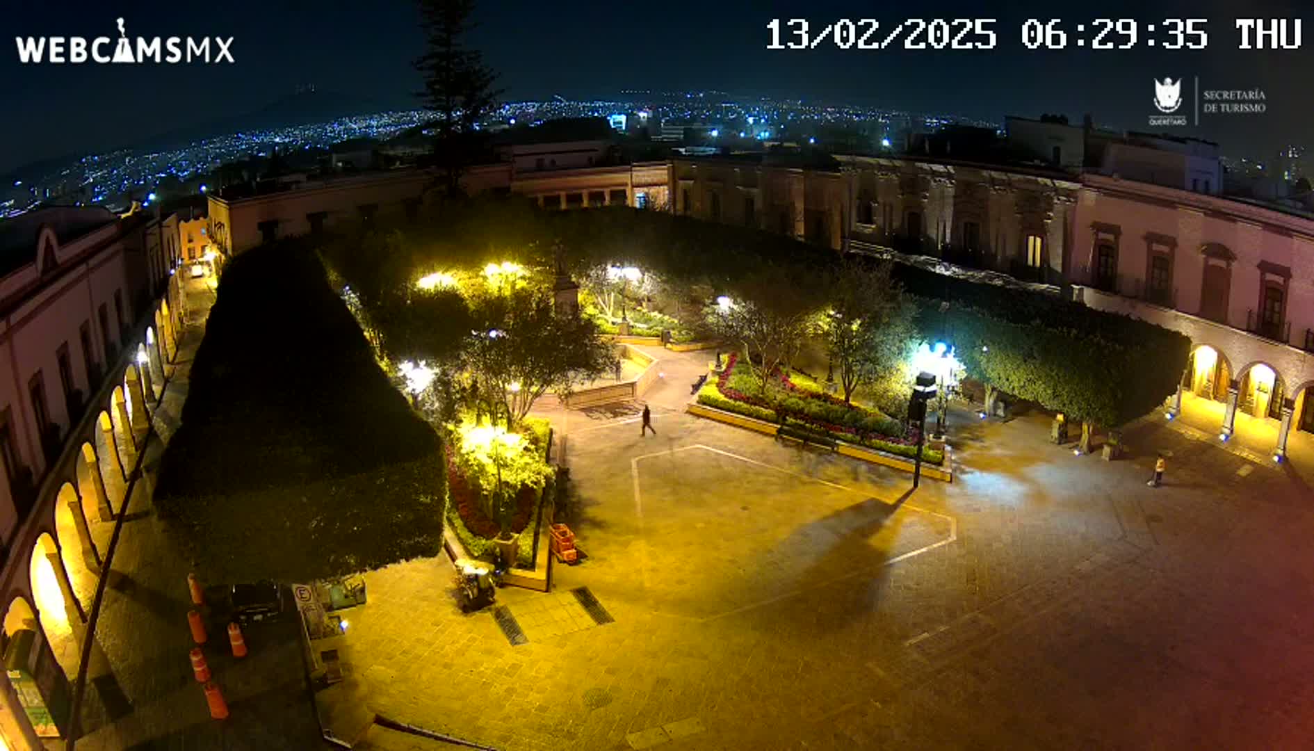 A nighttime aerial view of a brightly lit plaza surrounded by buildings, with a few pedestrians visible.