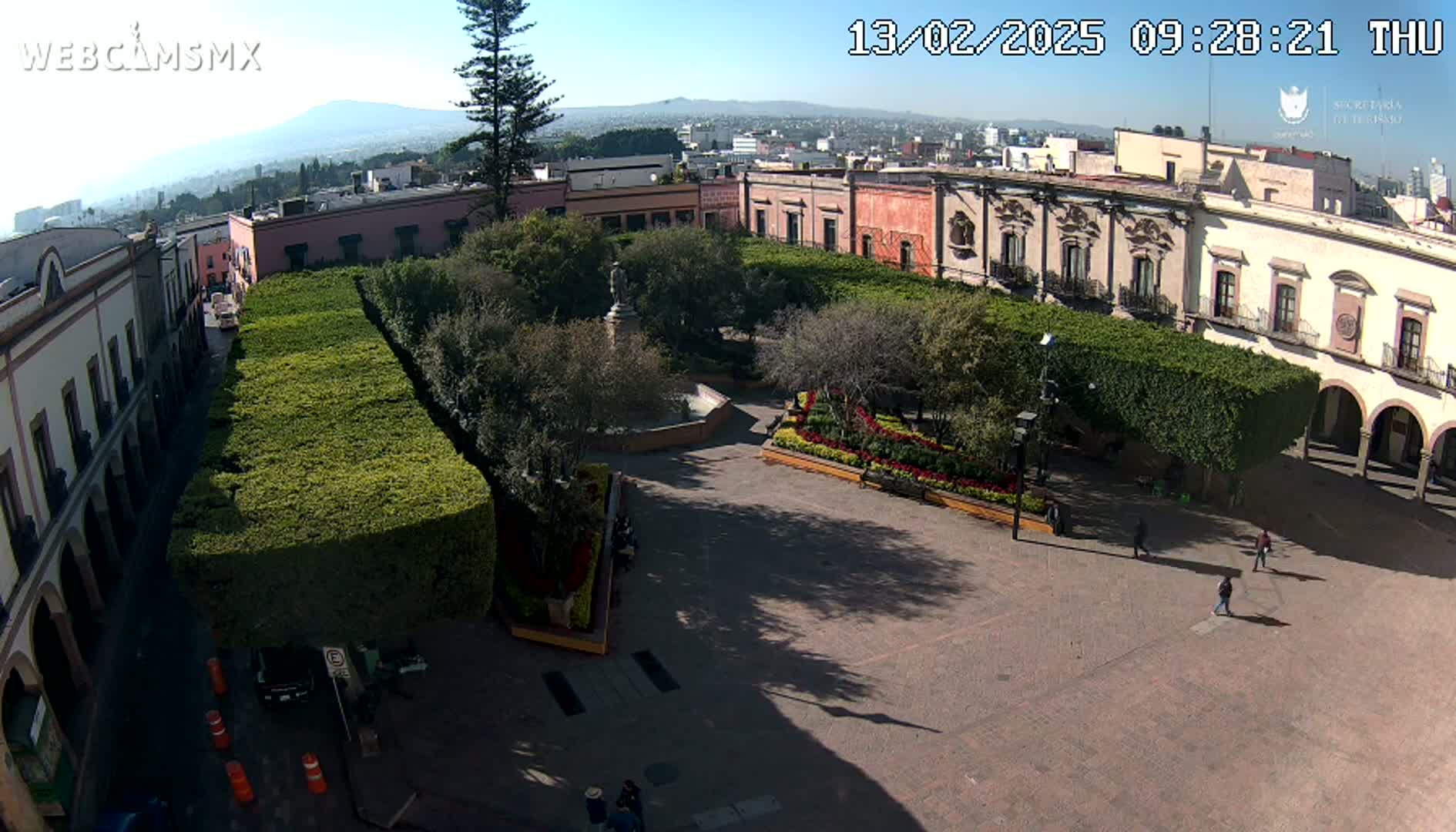 A sunny day shows an aerial view of a town square with neatly trimmed hedges, buildings, a fountain, and a few pedestrians.