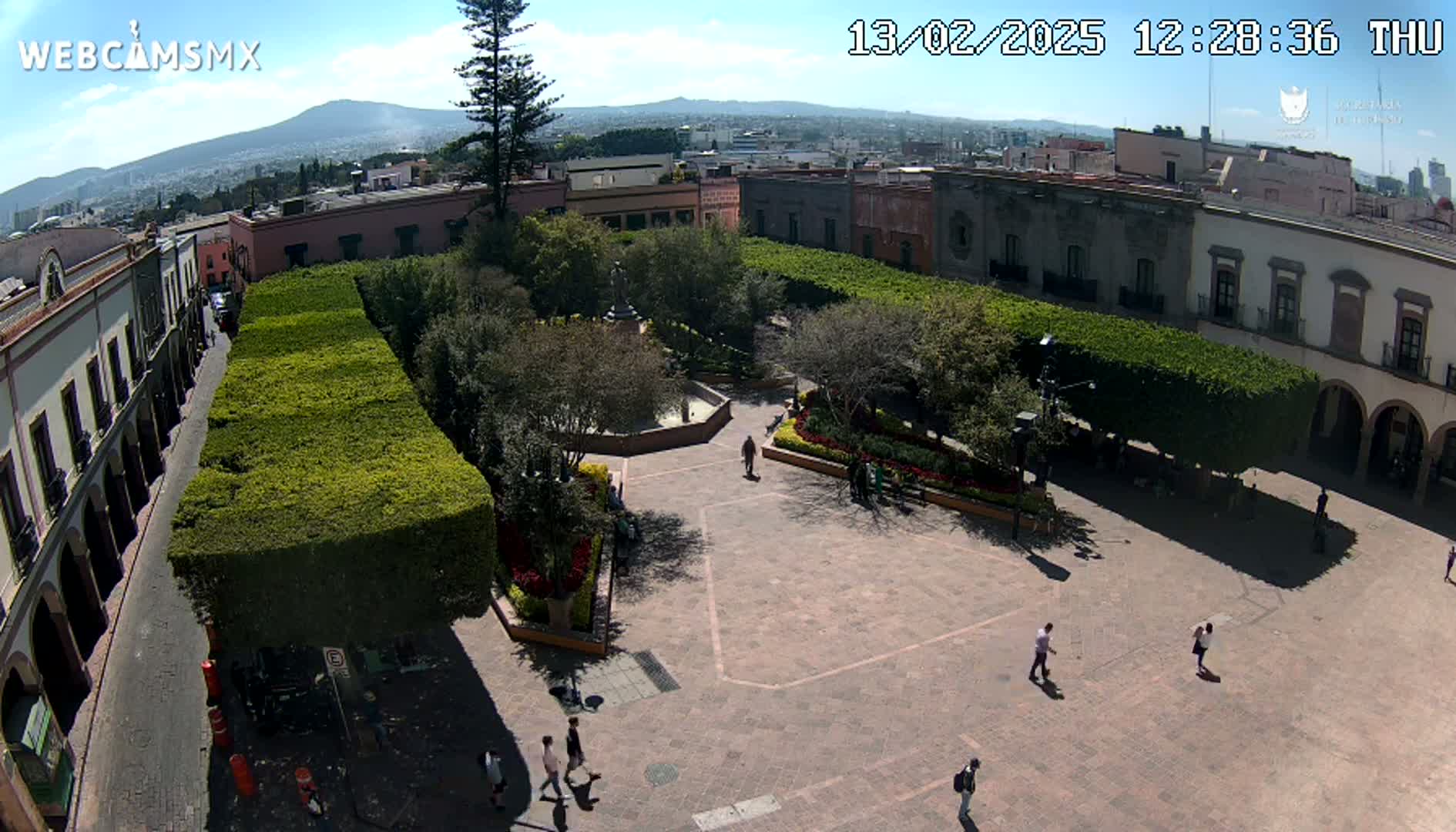 A high-angle view of a plaza, surrounded by buildings and trimmed hedges, on a sunny day with a mountain visible in the distance.