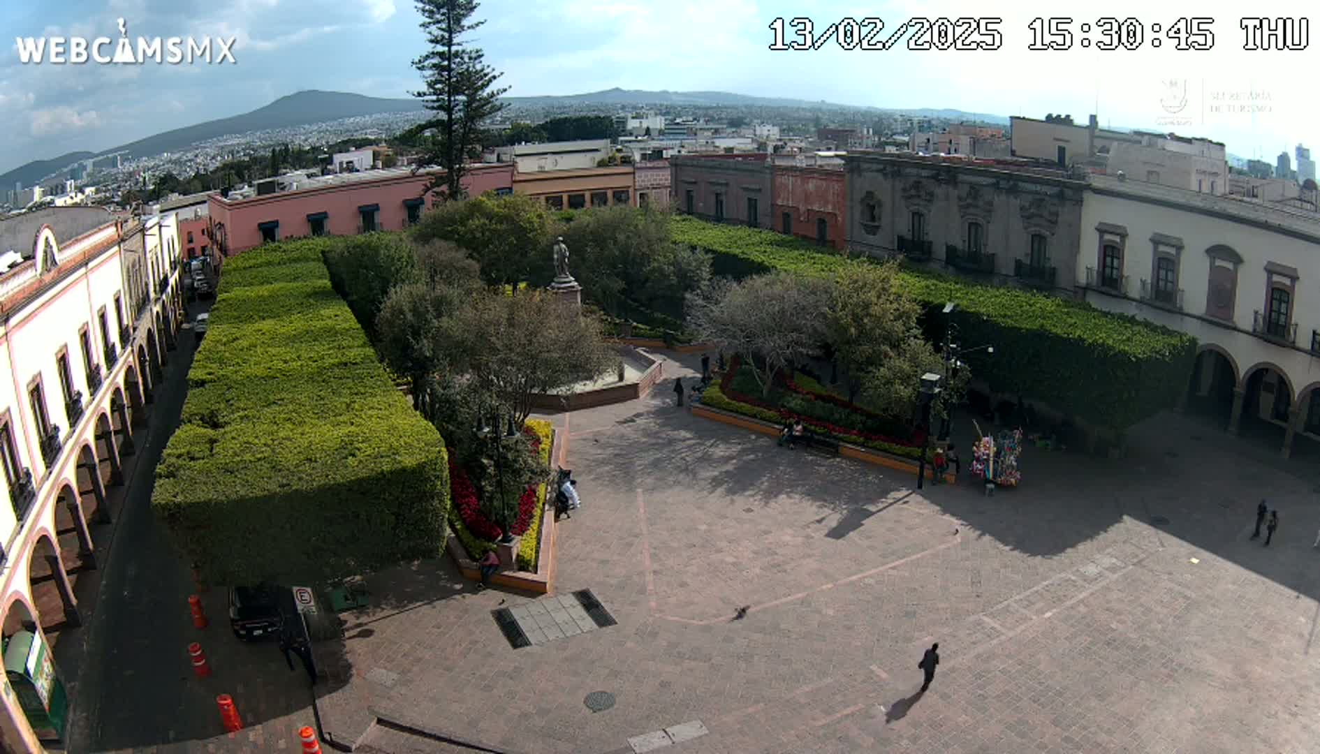 A sunny plaza with a statue at its center, surrounded by low hedges, colonial-style buildings, and a few people, is viewed from above.