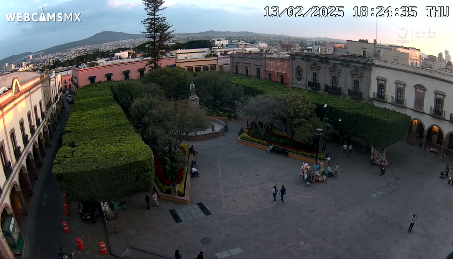 An aerial view of a town square at dusk, showing colonial-style buildings surrounding a plaza with neatly trimmed hedges, a central fountain, and a few people walking around.