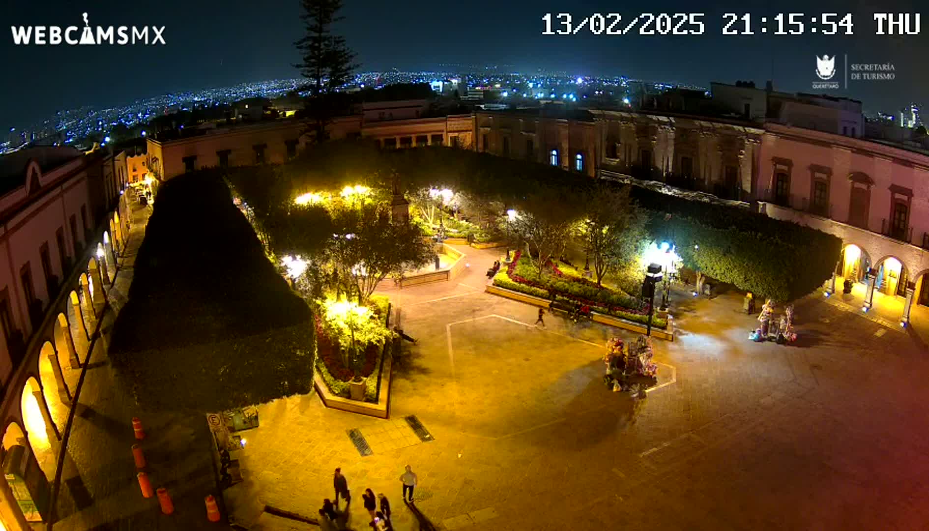 A nighttime, high-angle view of a brightly lit town square with surrounding buildings, landscaping, and a few pedestrians.