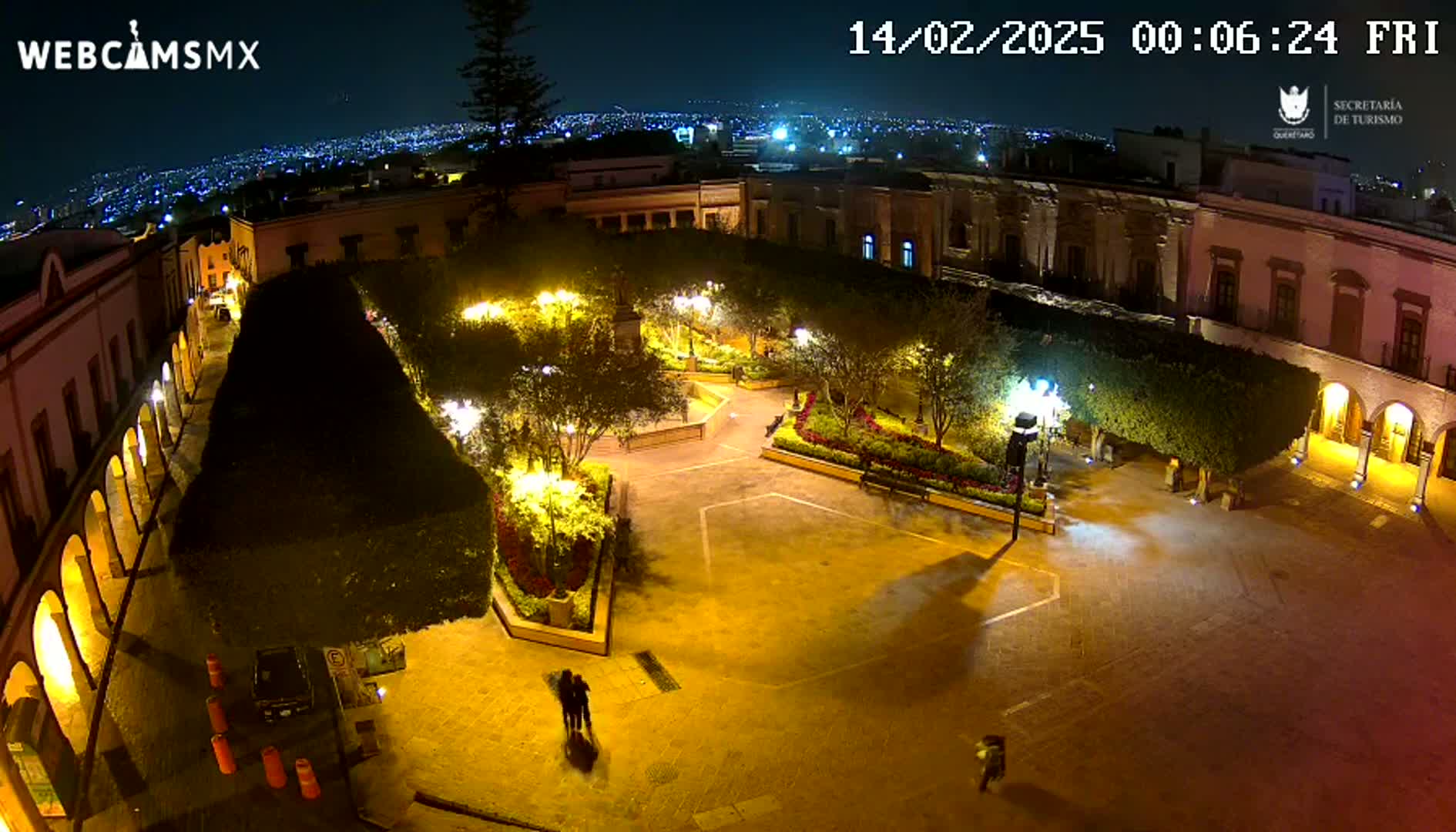 A nighttime overhead view of a brightly lit plaza surrounded by buildings, with a city skyline visible in the distance under a clear sky.