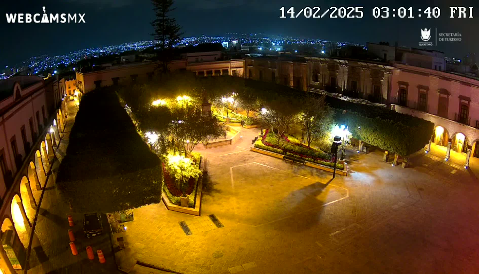 An illuminated plaza at night, surrounded by buildings, with a city skyline visible in the distance under a clear sky.