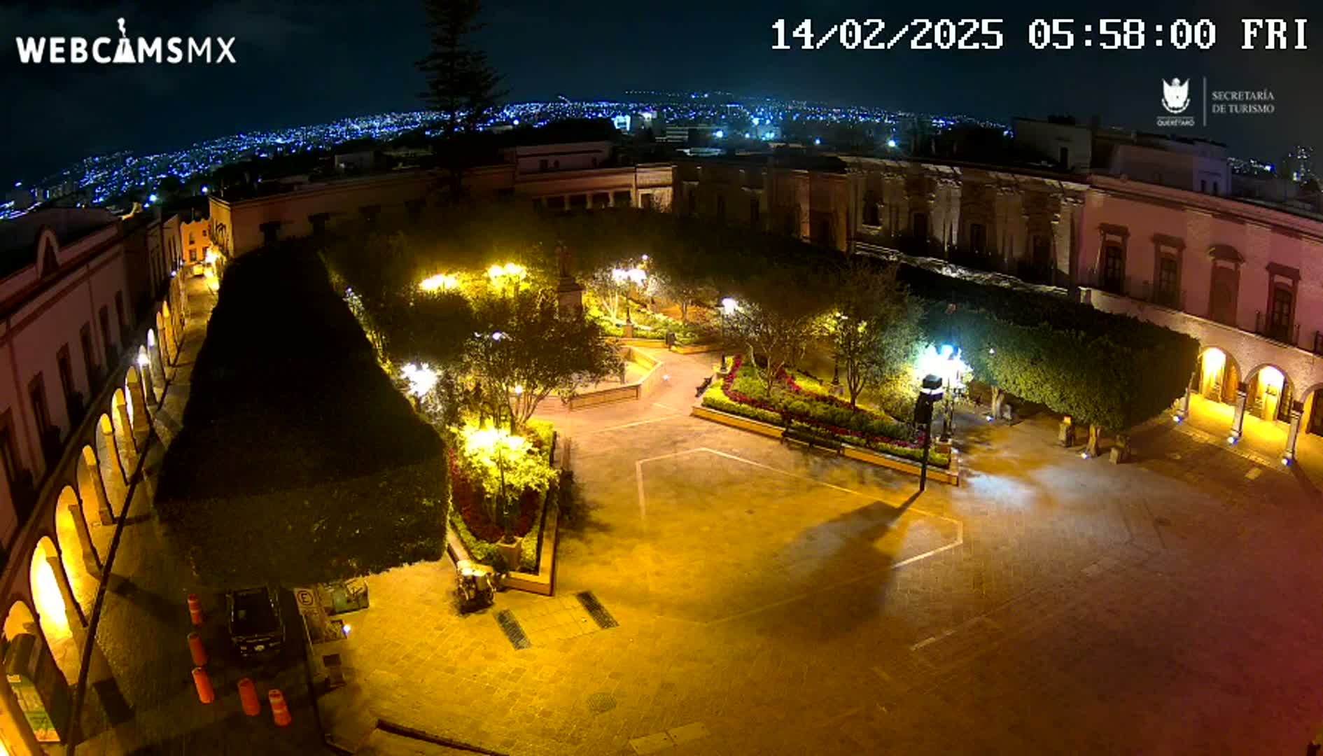 A brightly lit plaza at night, surrounded by colonial-style buildings, with a city skyline visible in the distance under a clear sky.